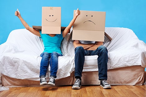 A cheerful woman and unhappy-looking man sit on a sofa  with cardboard boxes on their heads on which their faces have been drawn