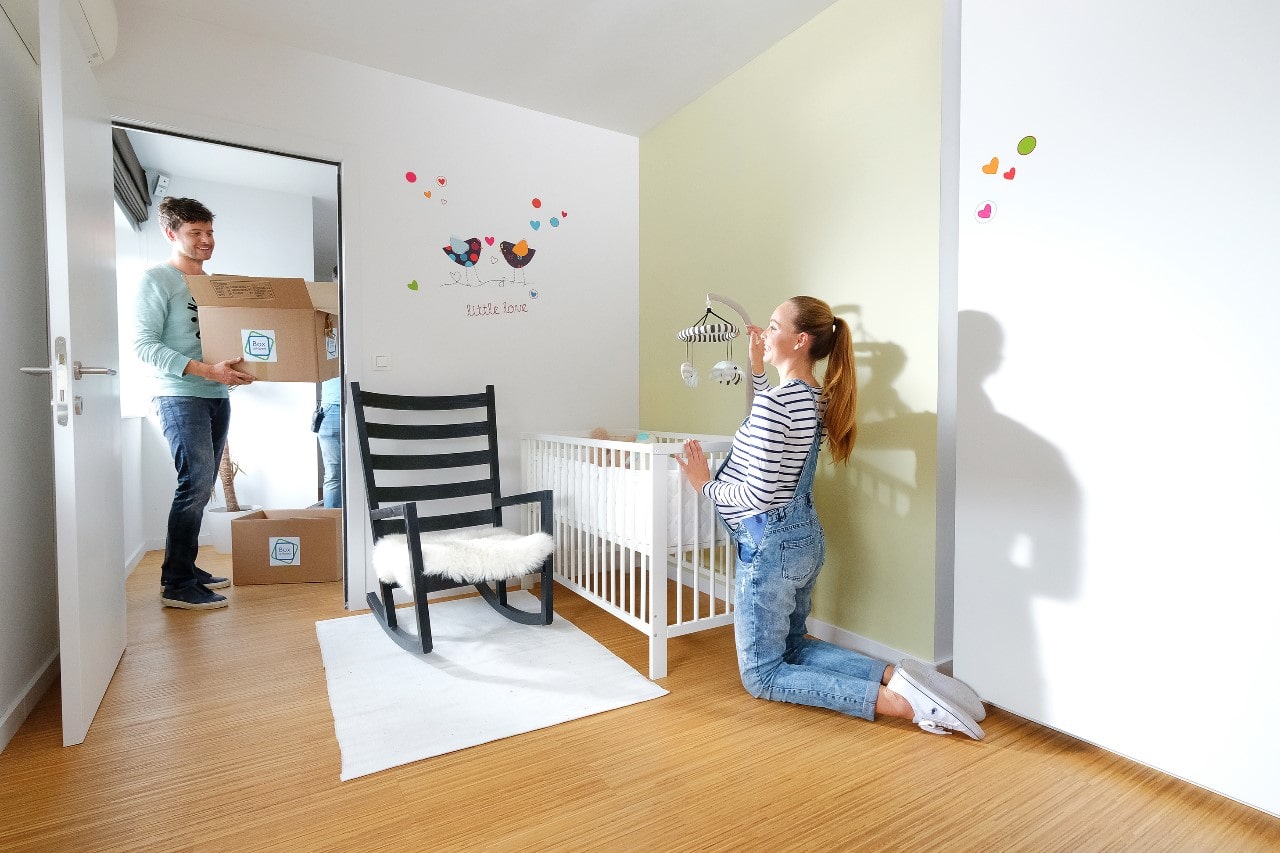 Un jeune homme se trouve dans la chambre d’enfant, et transporte une boîte de déménagement de Box@Home pendant que sa petite amie s’occupe du faire le lit de bébé.