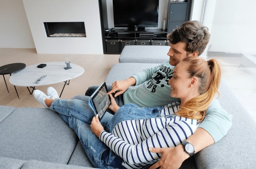 Les deux personnes de ce jeune couple sont assises dans le fauteuil. À l'aide de leur tablette, ils commandent leur espace d’entreposage en ligne sur le site Internet de Box@Home.
