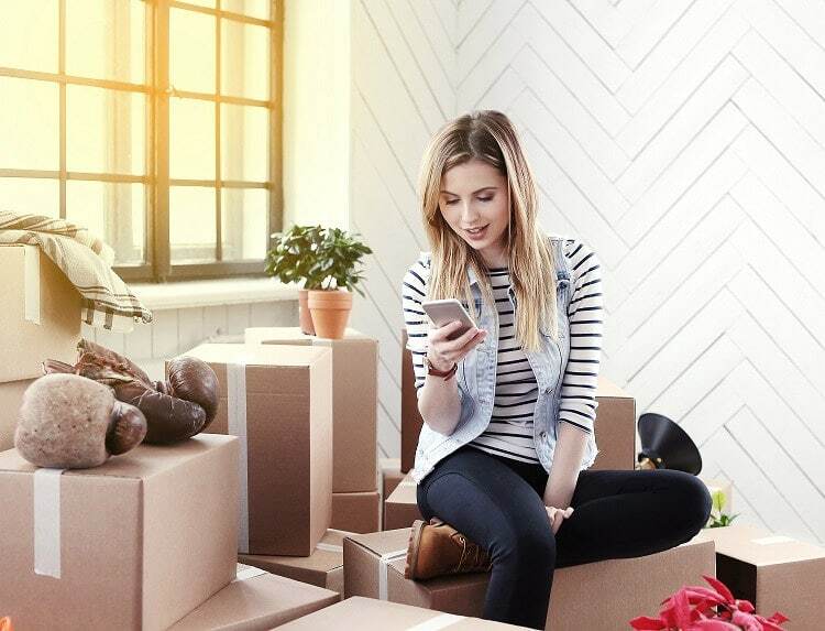 Une étudiante vivant en kot est assise sur une pile de boîtes de déménagement, et commande son espace d’entreposage en ligne sur le site Internet de Box@Home.